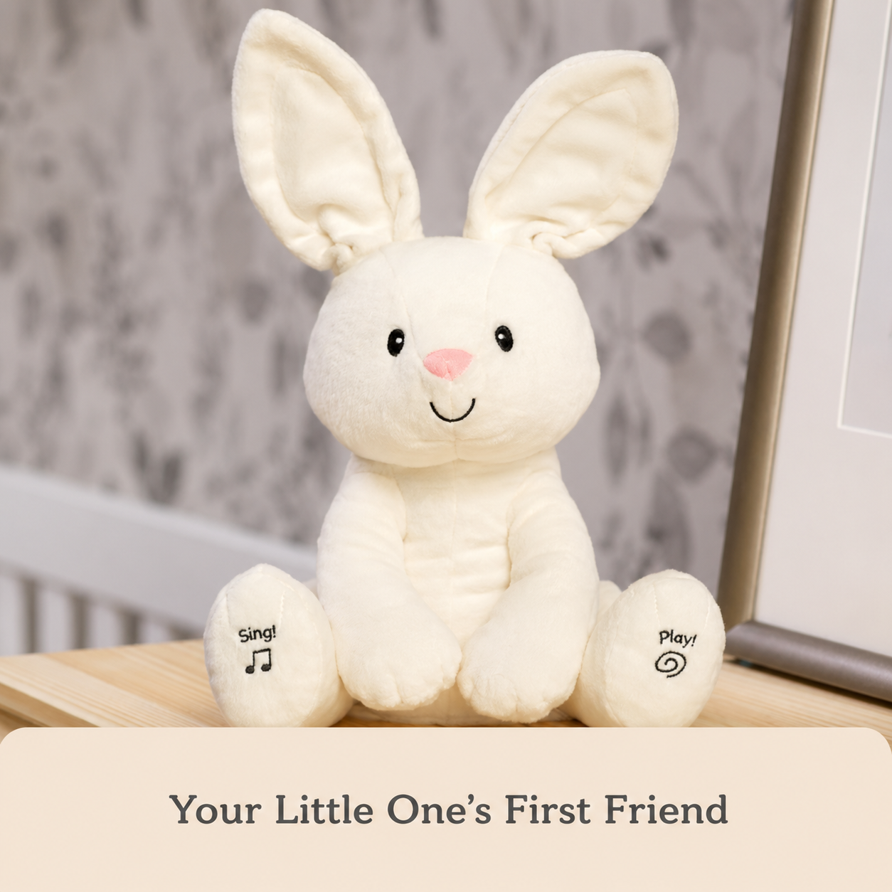 A cream-colored plush bunny toy sitting on a wooden surface, with printed text on its feet reading 'sing!' and 'play!'. A beige banner at the bottom reads 'Your Little One's First Friend'.