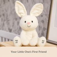 A cream-colored plush bunny toy sitting on a wooden surface, with printed text on its feet reading 'sing!' and 'play!'. A beige banner at the bottom reads 'Your Little One's First Friend'.