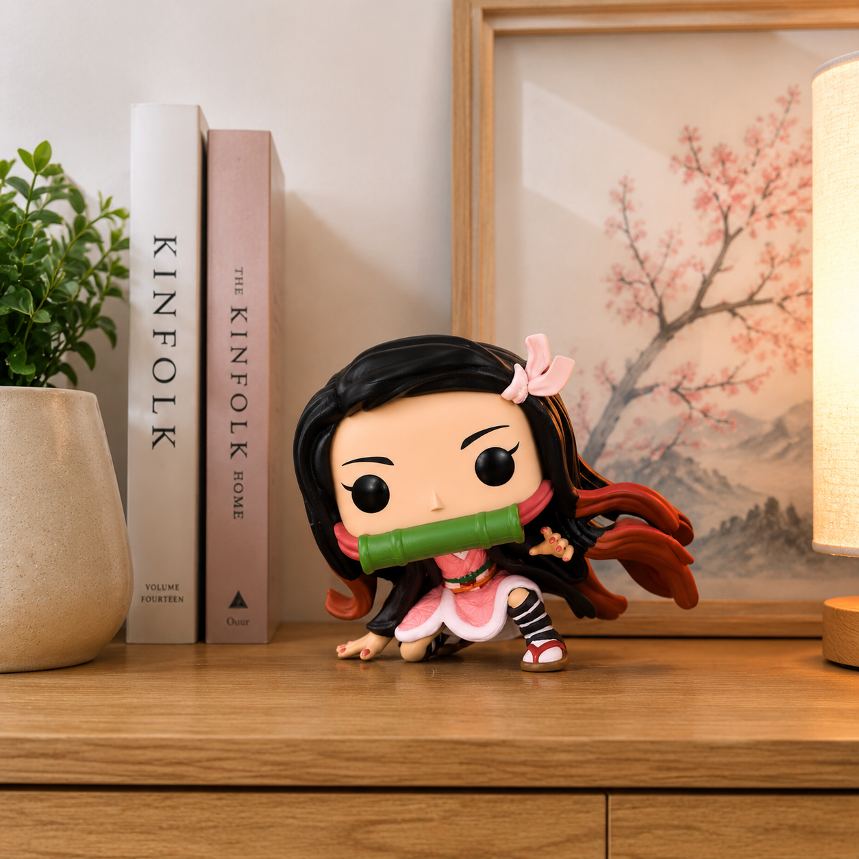 a pink bow, and a green object in its mouth, displayed on a wooden surface beside books, a plant, and a lit lamp.