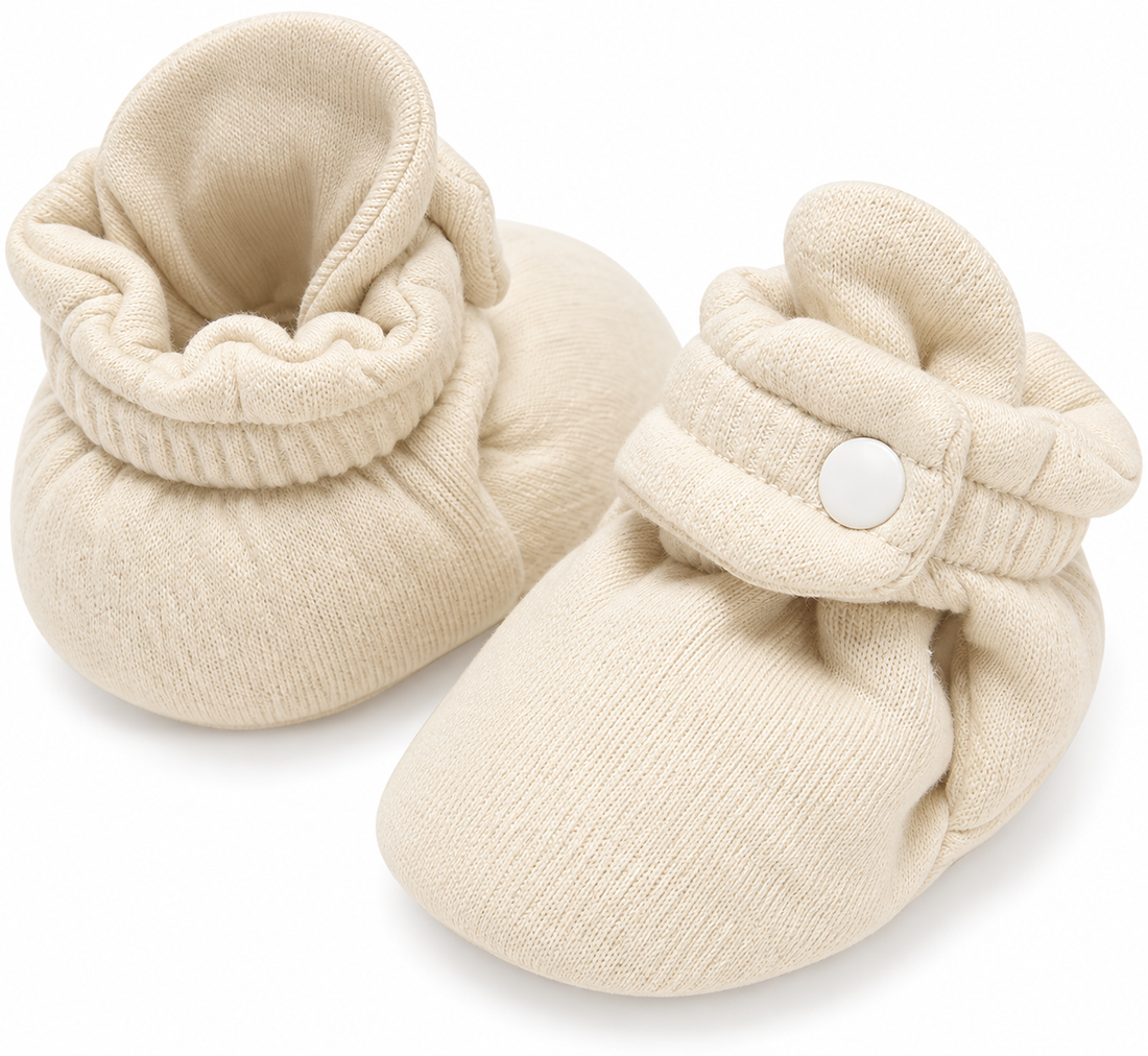 Pair of cream knitted baby booties with folded ankle cuffs and snap-button straps on a white background.