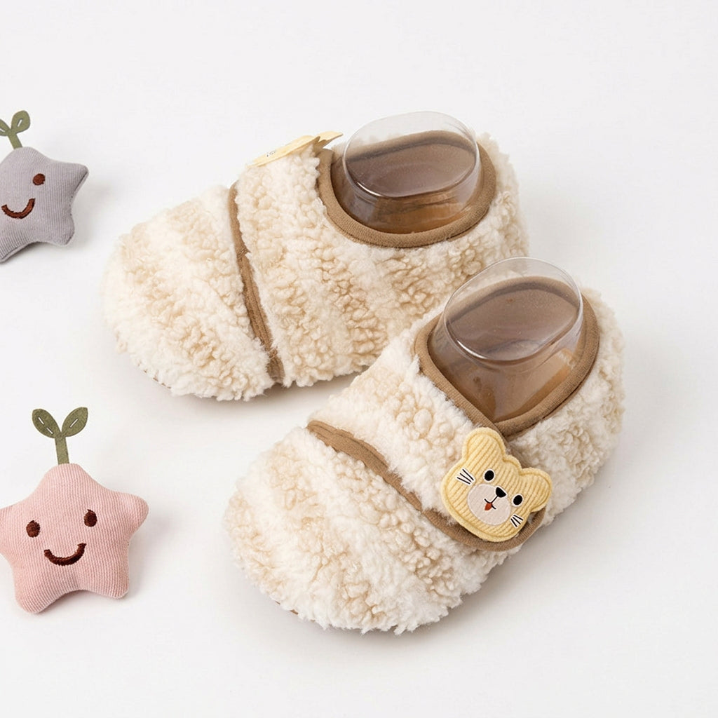 Pair of cream fleece baby booties with beige trim and a cute teddy bear patch on the strap.