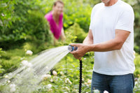 Close-up of a person holding a black-and-green garden hose nozzle, releasing a powerful stream of water across a vibrant garden.
